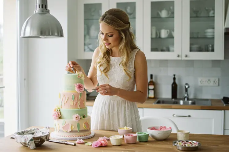 Bride in a cozy modern kitchen baking and decorating a wedding cake.