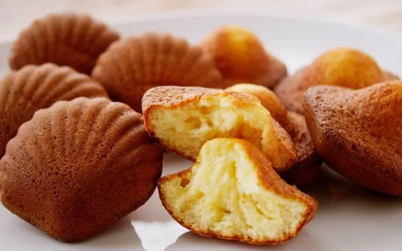 French madeleines, shell-shaped cakes, golden edges, rustic bakery table, soft natural light, cozy aesthetic.